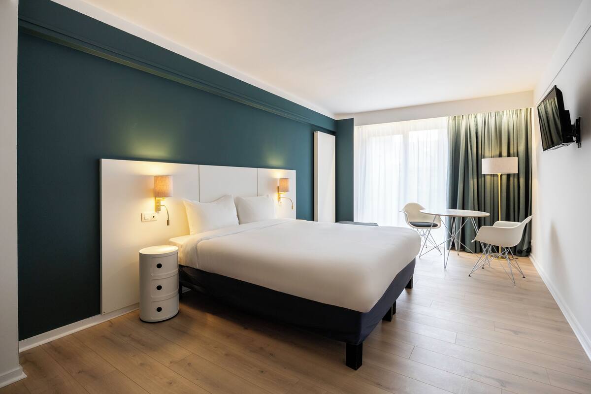 A modern hotel room with a large bed, bedside lamps, a small table with chairs, and a TV mounted on the wall.