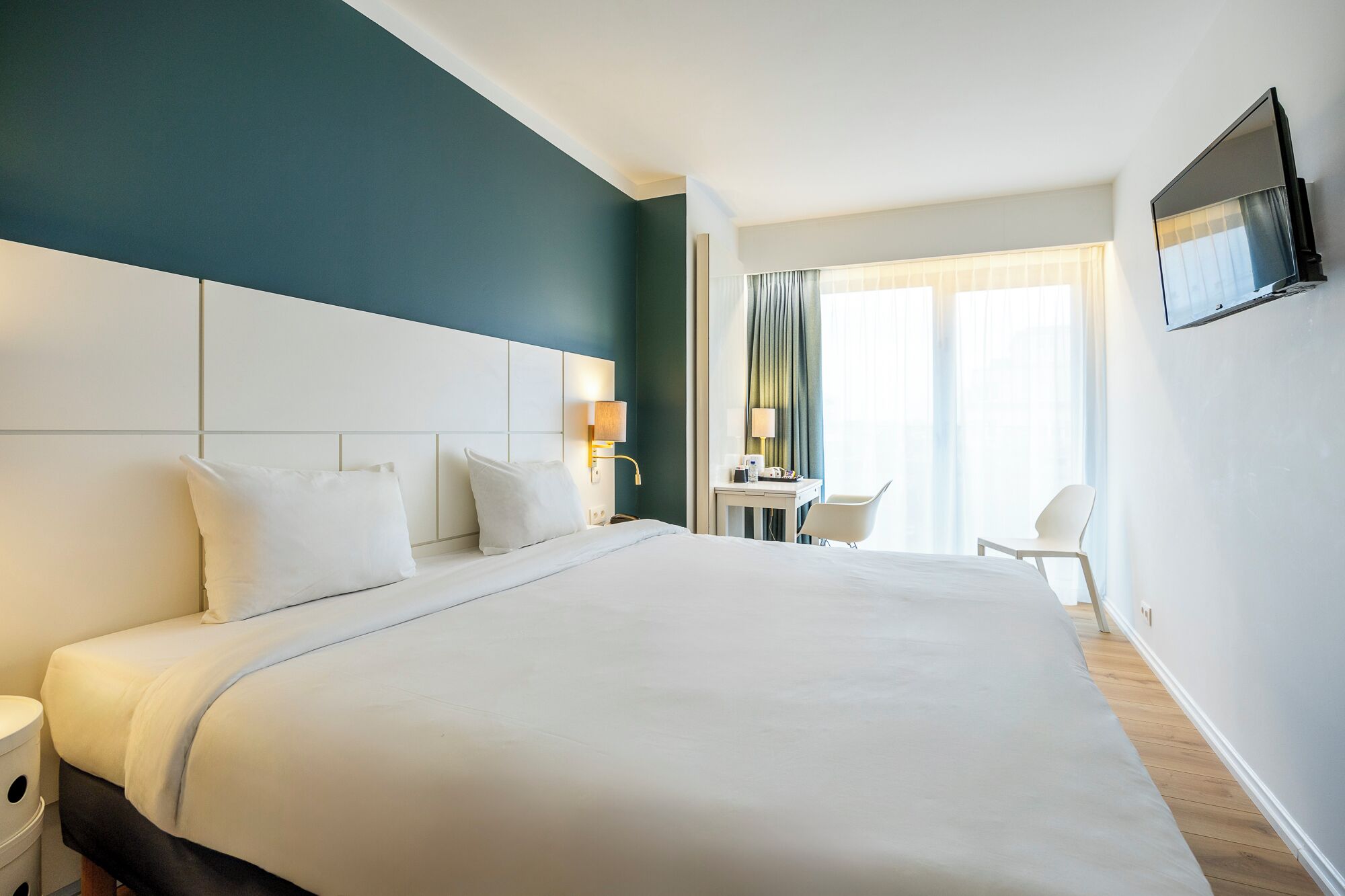 Ein Hotelzimmer mit einem großen Bett, Nachttischen, einem Schreibtisch mit Stuhl, einem an der Wand befestigten Fernseher und einem Fenster mit Vorhängen.