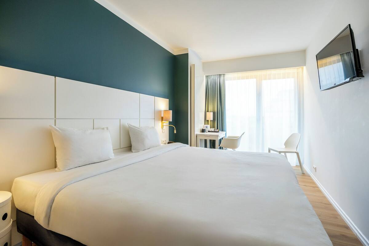A hotel room with a large bed, bedside tables, a desk with a chair, a TV mounted on the wall, and a window with curtains.
