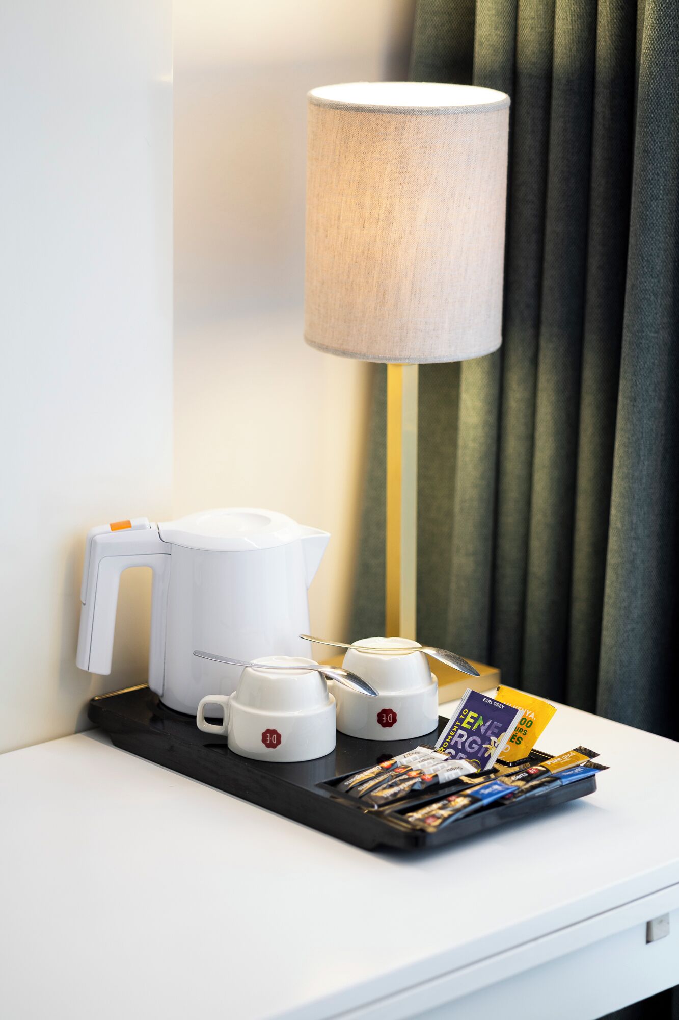 Ein Hotelzimmer mit einem weißen Nachttisch, einem weißen Wasserkocher, zwei weißen Tassen, einer Ablage mit Zeitschriften und einer Lampe.