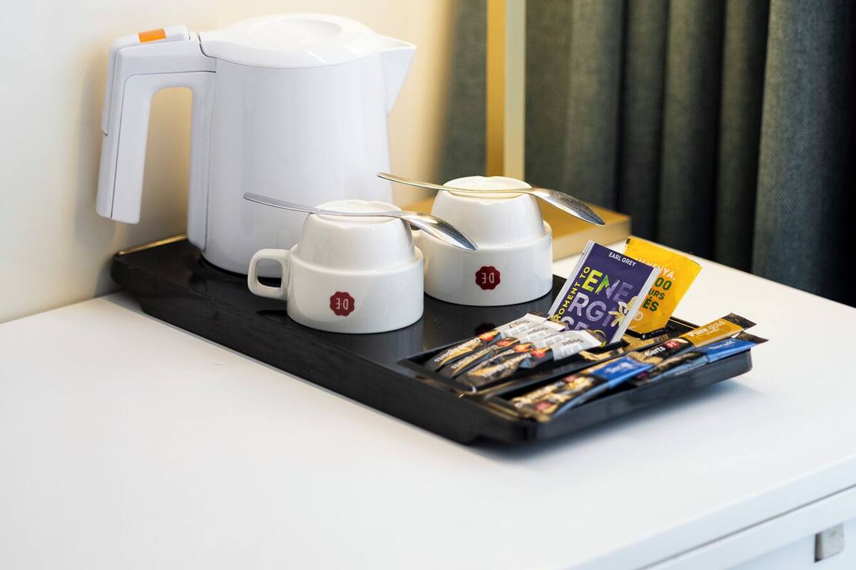 A hotel room with a white bedside table, a white electric kettle, two white cups, a tray with magazines, and a lamp.
