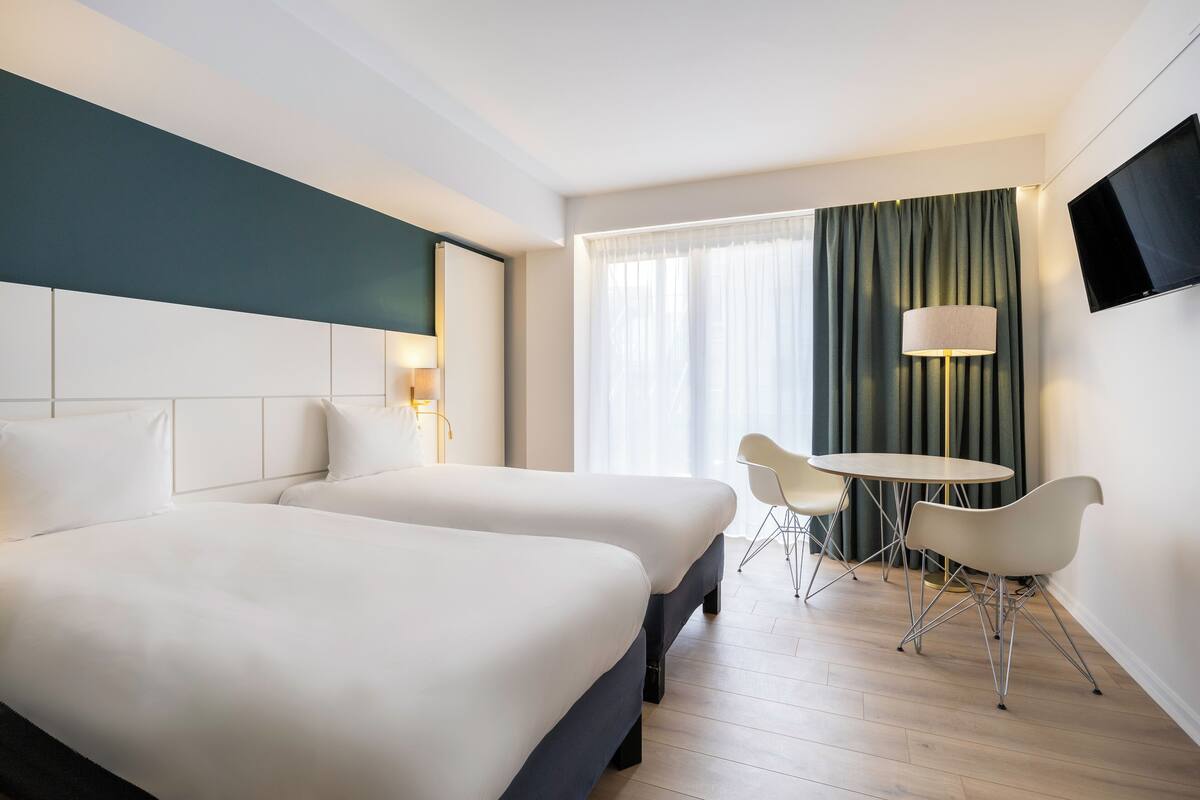A hotel room with two beds, a desk, a chair, a TV, and a window with curtains.
