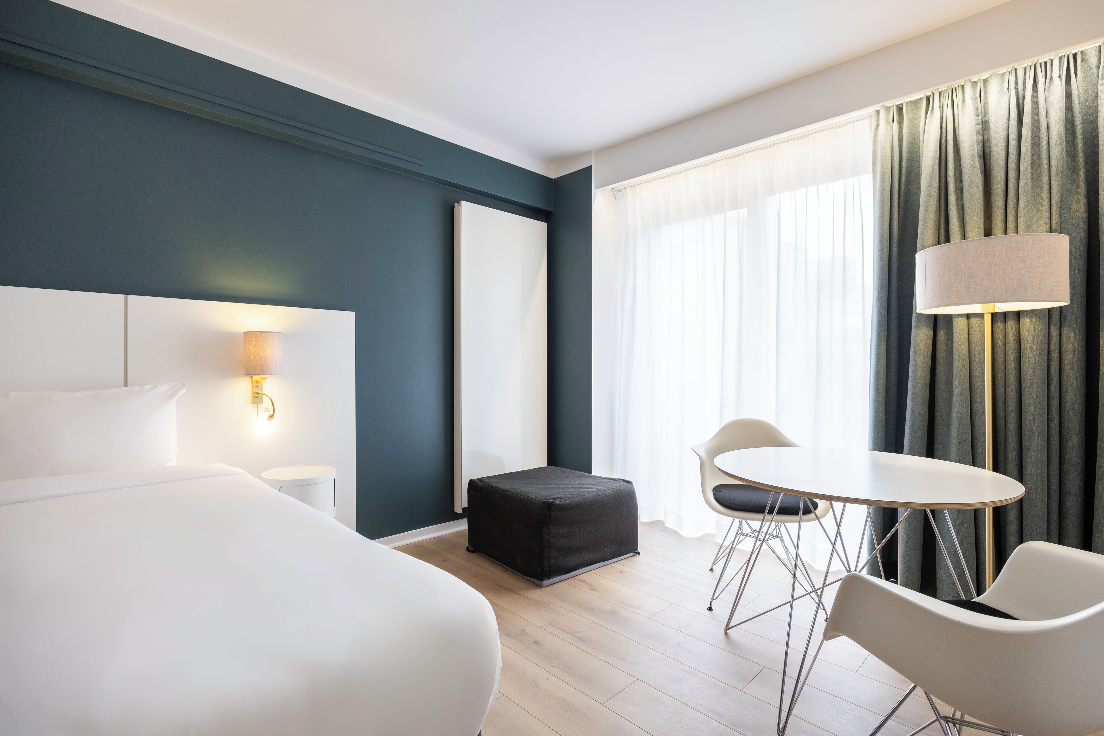 Una camera d'albergo moderna con un letto grande, un tavolino con sedie, una lampada e vista all'aperto attraverso una finestra con tende.