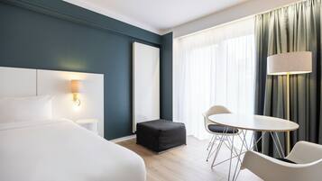 Una camera d'albergo moderna con un letto grande, un tavolino con sedie, una lampada e vista all'aperto attraverso una finestra con tende.
