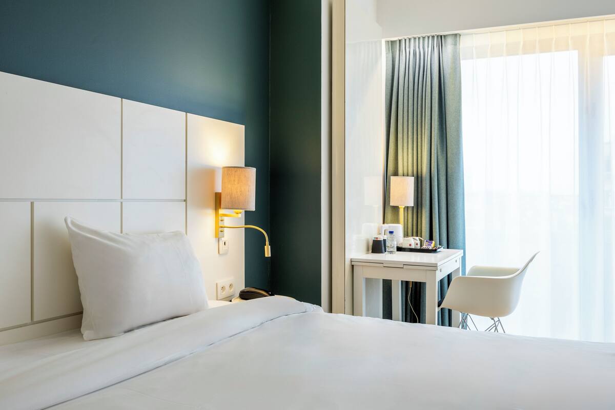 A hotel room with a bed, bedside table, lamp, chair, and a window with curtains.