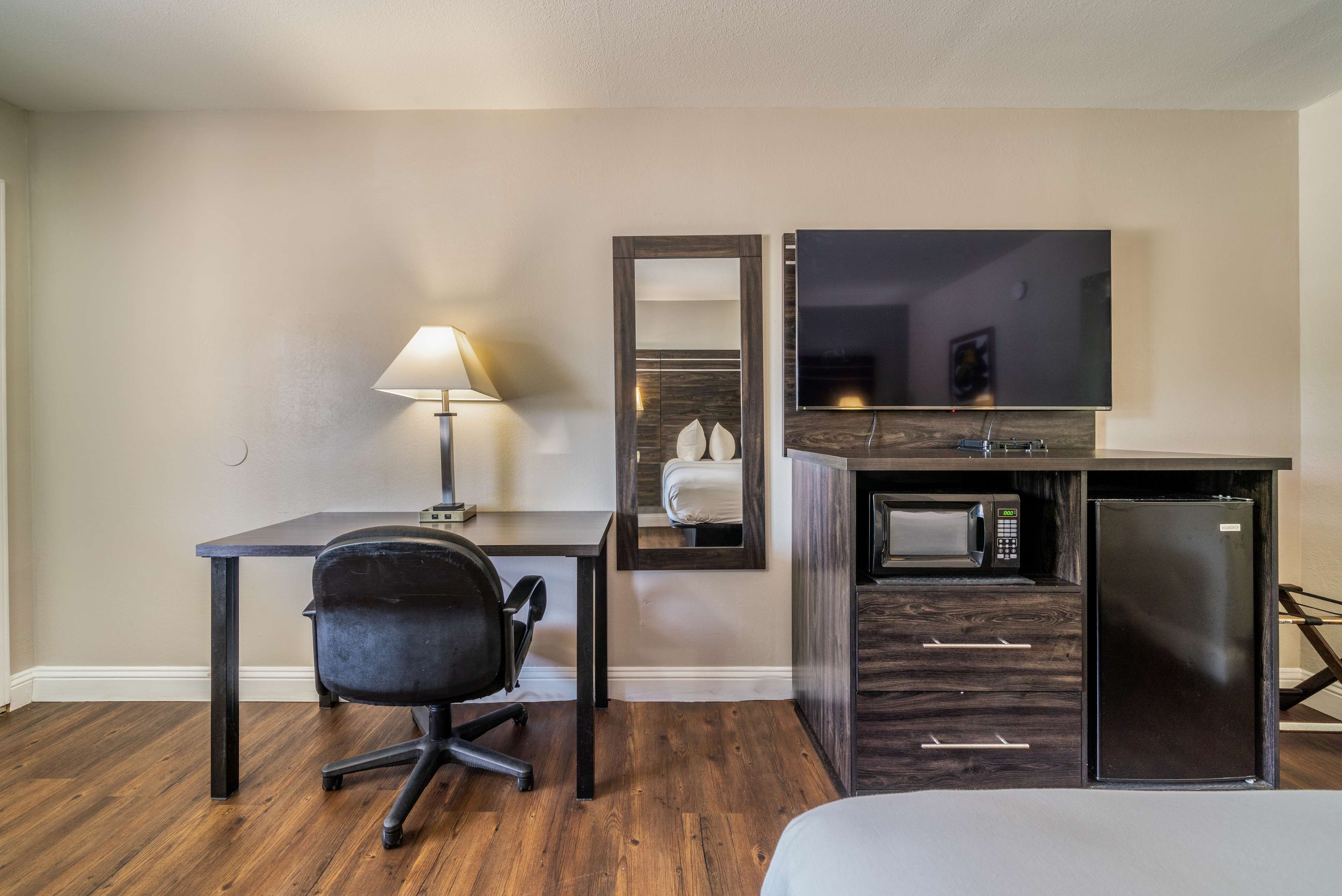 A compact hotel room with a wooden floor, a desk with a chair, a small kitchenette with a microwave and refrigerator, a bedside table, and a large mirror.