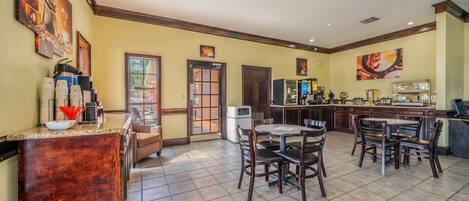 A spacious dining area with a central round table and chairs, a coffee station with multiple cups, and a kitchen area in the background.
