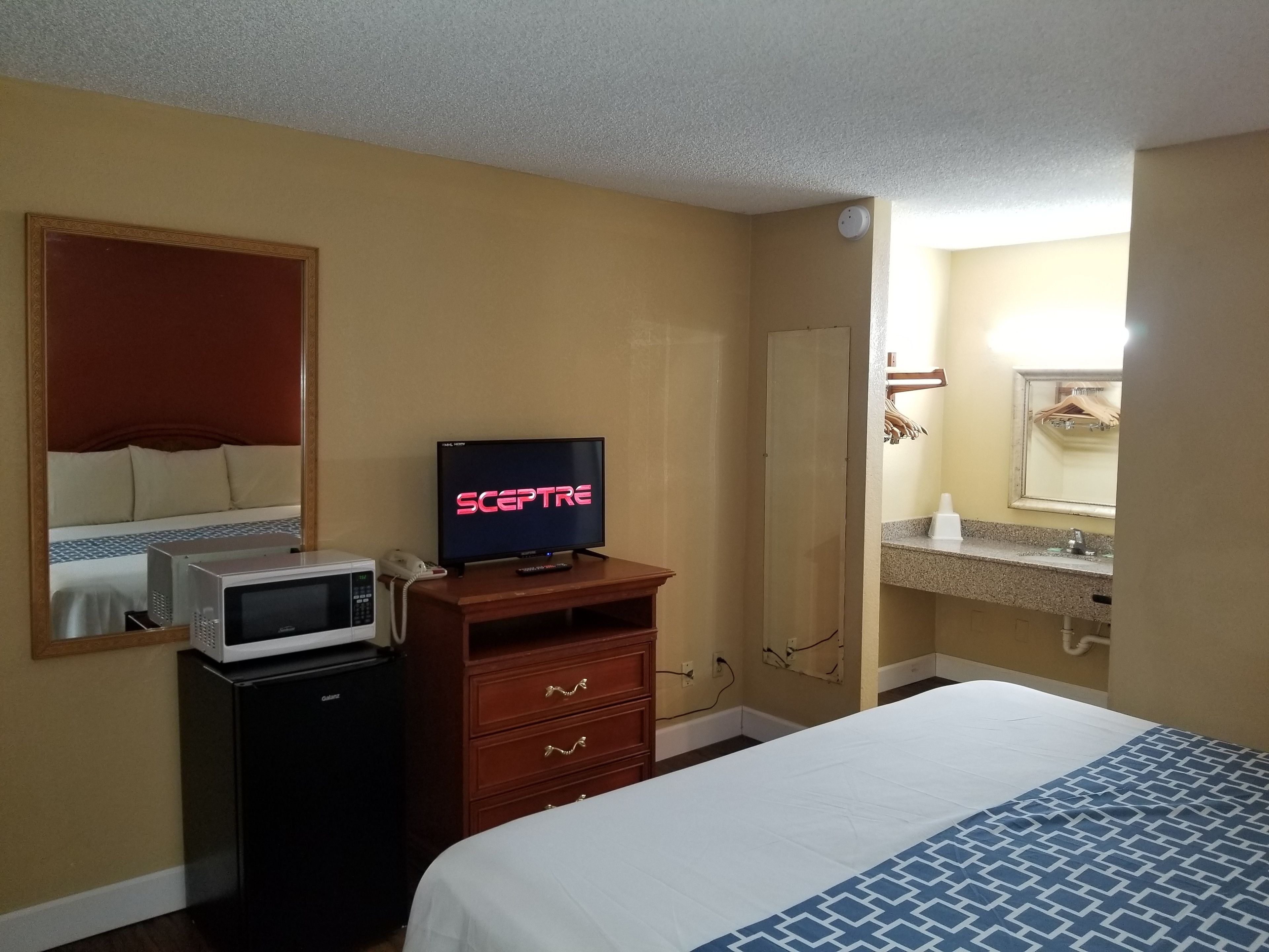 Une chambre d’hôtel comprenant un lit, un téléviseur diffusant l’émission « SCEPTRE », un four à micro-ondes, un petit meuble, un miroir et une salle de bain avec un lavabo et un porte-serviettes.