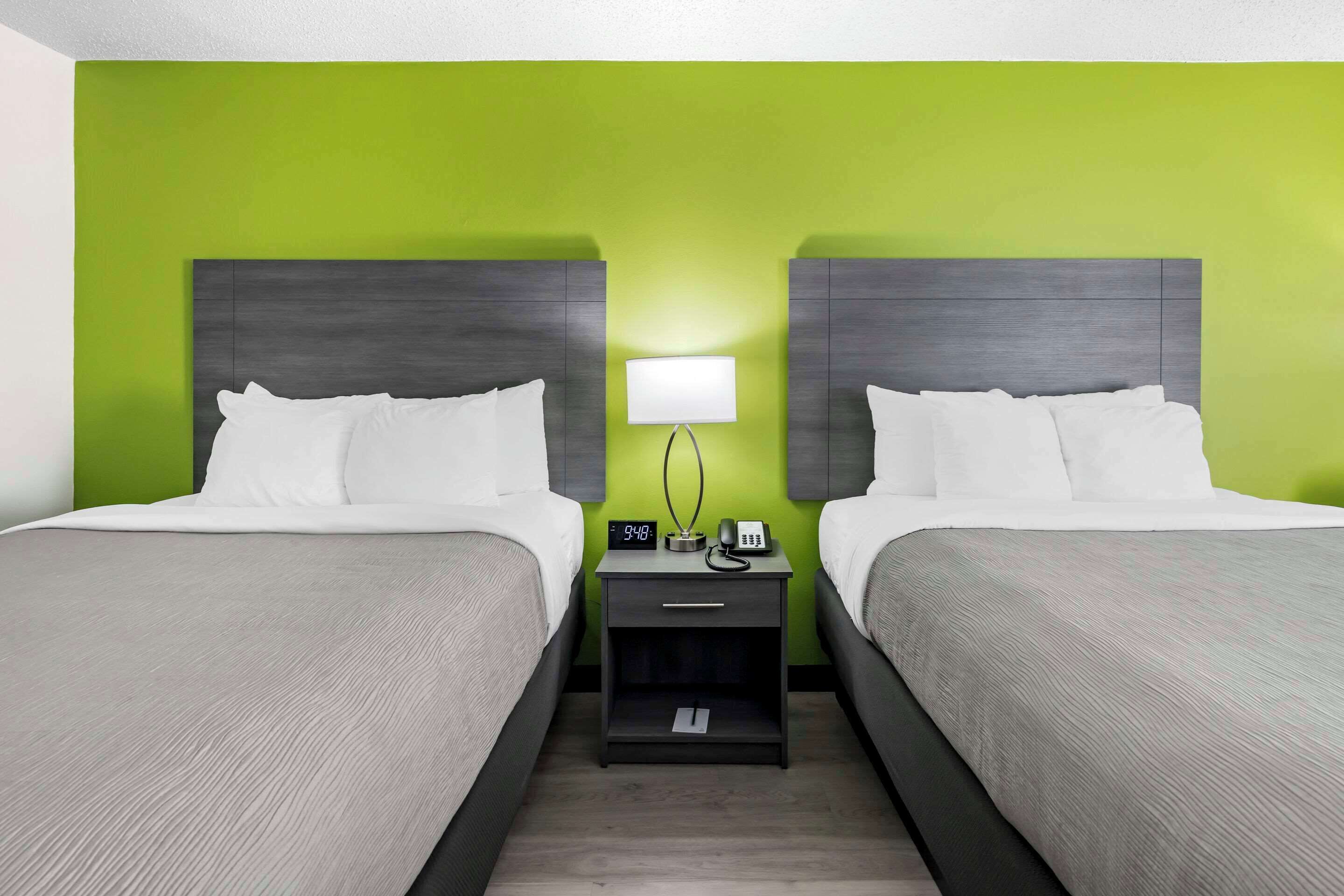 A hotel room with two beds, a nightstand with a lamp, a clock, and a phone.