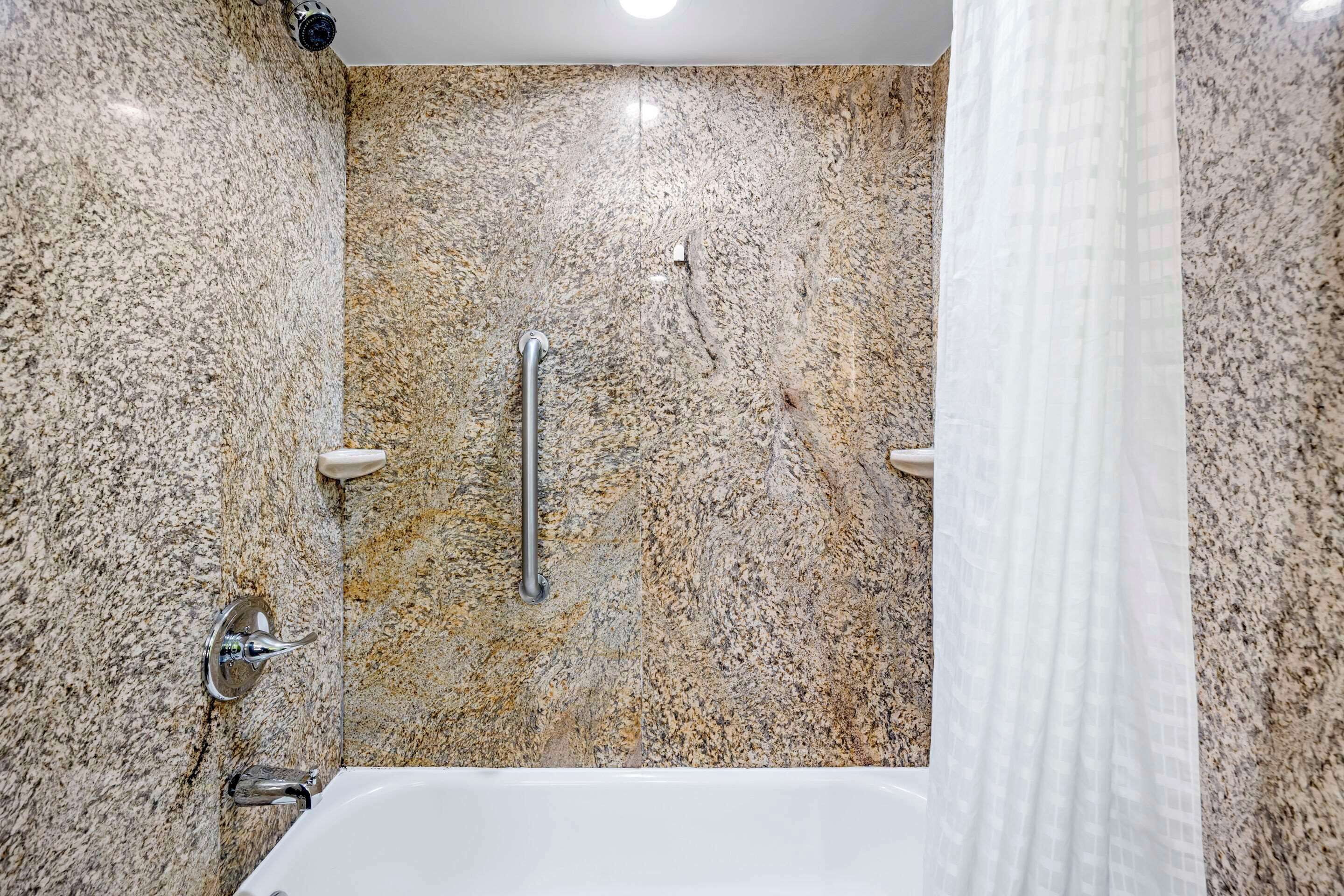 A bathroom with a granite countertop, a white bathtub, and a handheld showerhead.