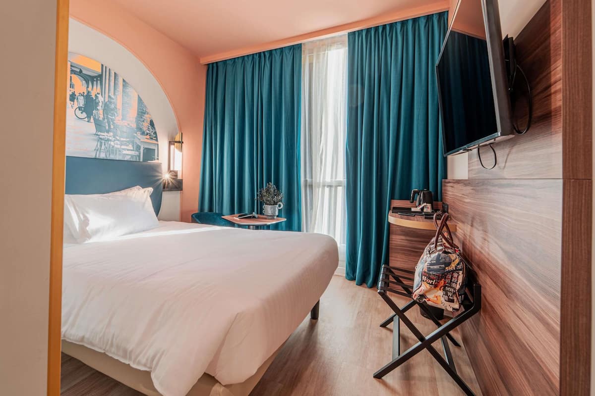 Ein Hotelzimmer mit Bett, Schreibtisch mit Stuhl, Fernseher und Fenster mit Vorhängen.