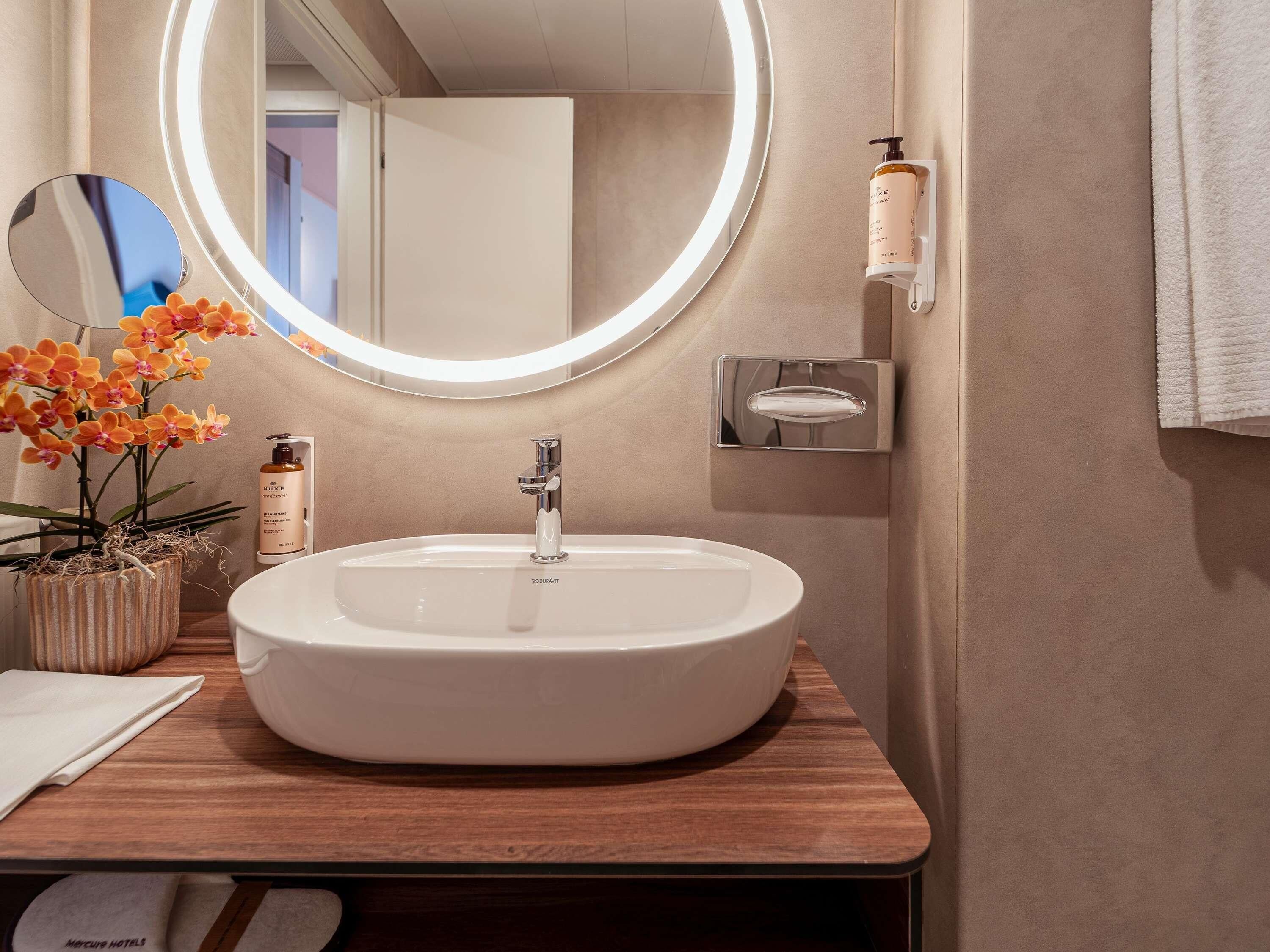 Ein Badezimmer mit einem weißen Waschbecken, einem runden Spiegel, einem Topf mit einer Orchidee und einer Shampoo-Flasche.