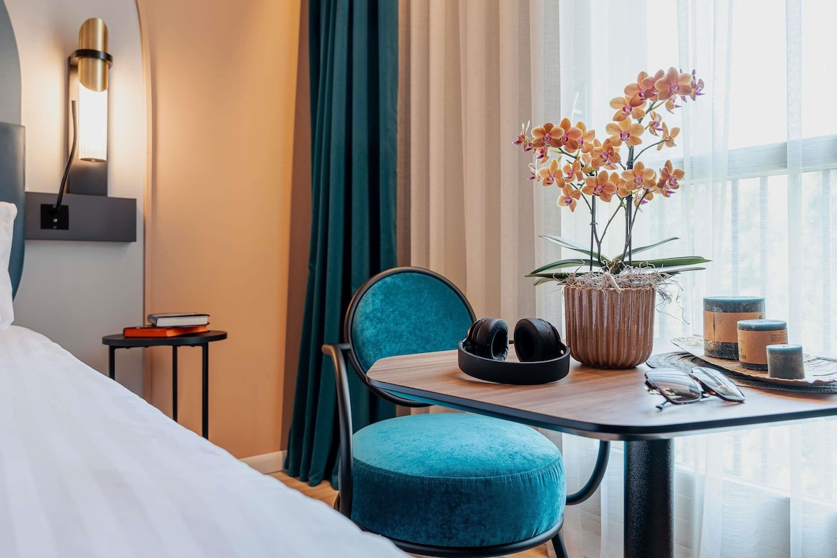 Une chambre d’hôtel avec un lit, un bureau et une chaise, une orchidée en pot et une fenêtre avec des rideaux.
