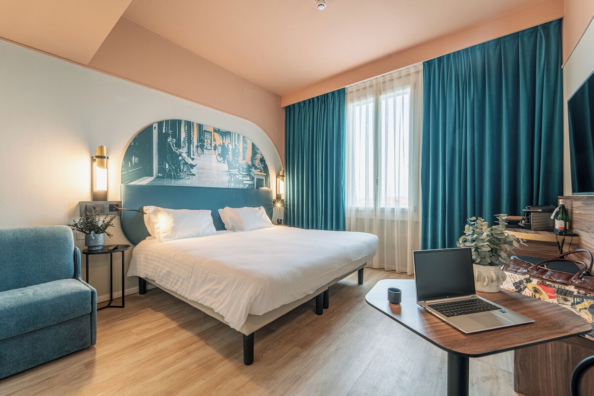 A modern hotel room with a bed, a desk with a laptop, a chair, a nightstand with a lamp, and a large window with curtains.