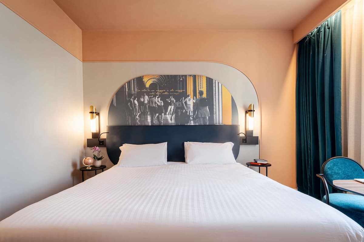 A hotel room with a large bed, two bedside tables, a headboard with a mural, and a chair.