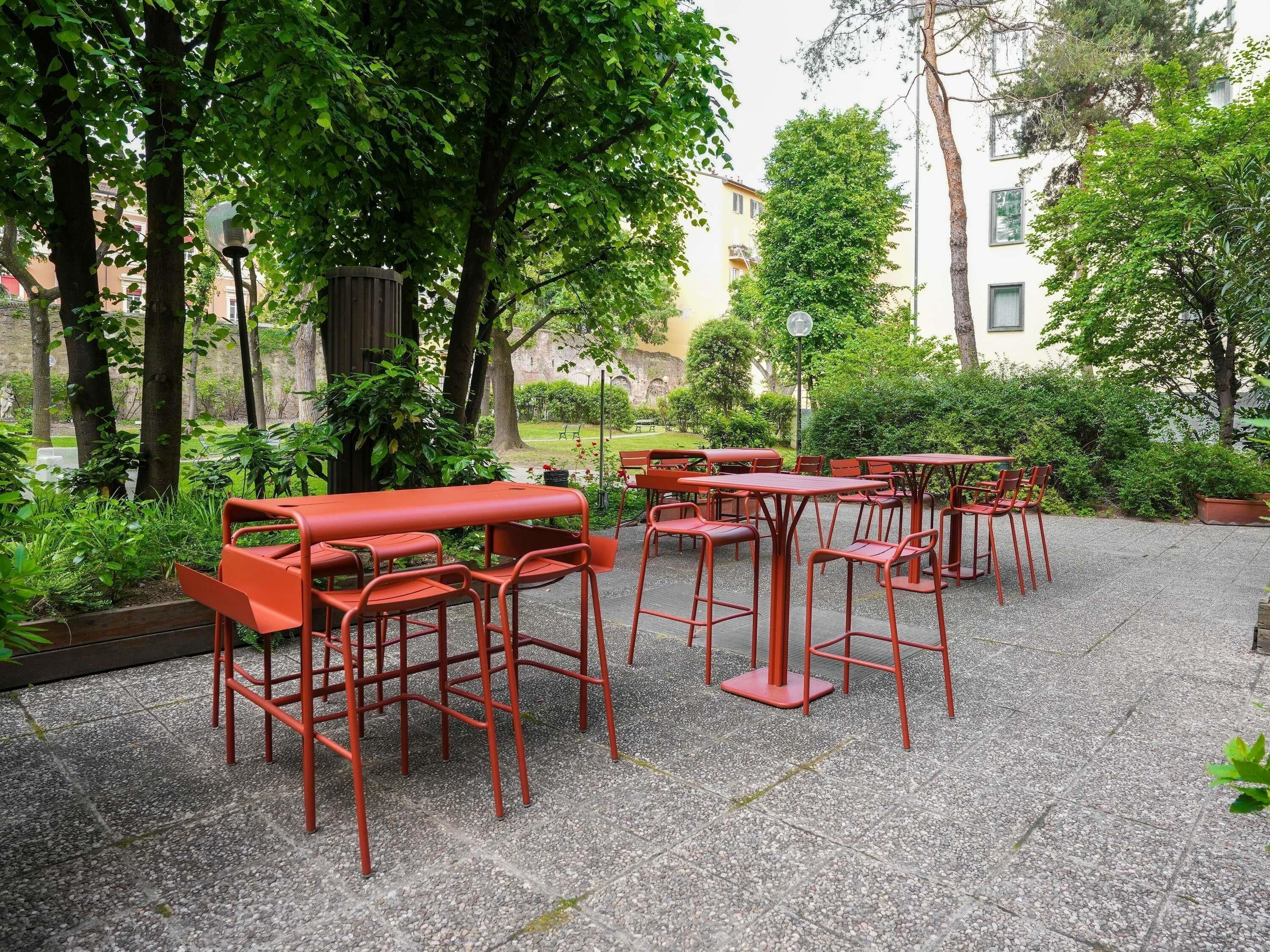 Un espacio al aire libre con mesas y sillas de metal rojo, rodeado de follaje y árboles.