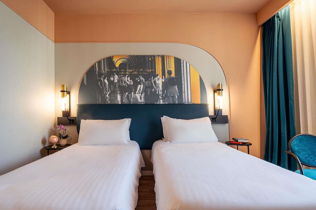 Ein Hotelzimmer mit zwei Betten, einem großen Kopfteil mit Wandgemälde, Nachttischlampen, einem kleinen Tisch mit einem Buch und einem Sessel.