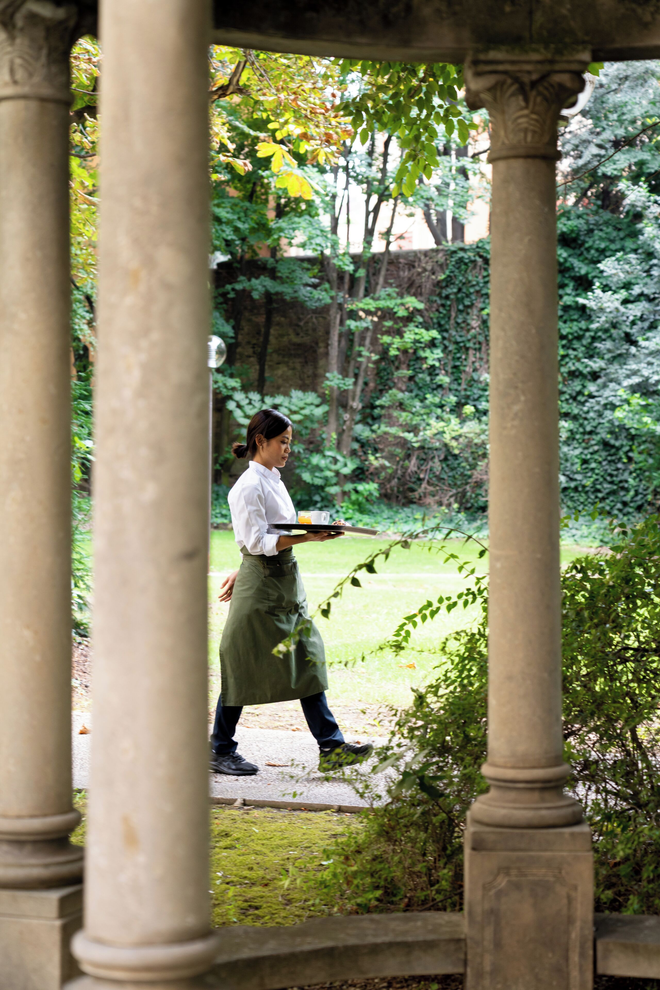 Eine Person geht durch einen Garten mit Stein-Säulen und viel Grün.