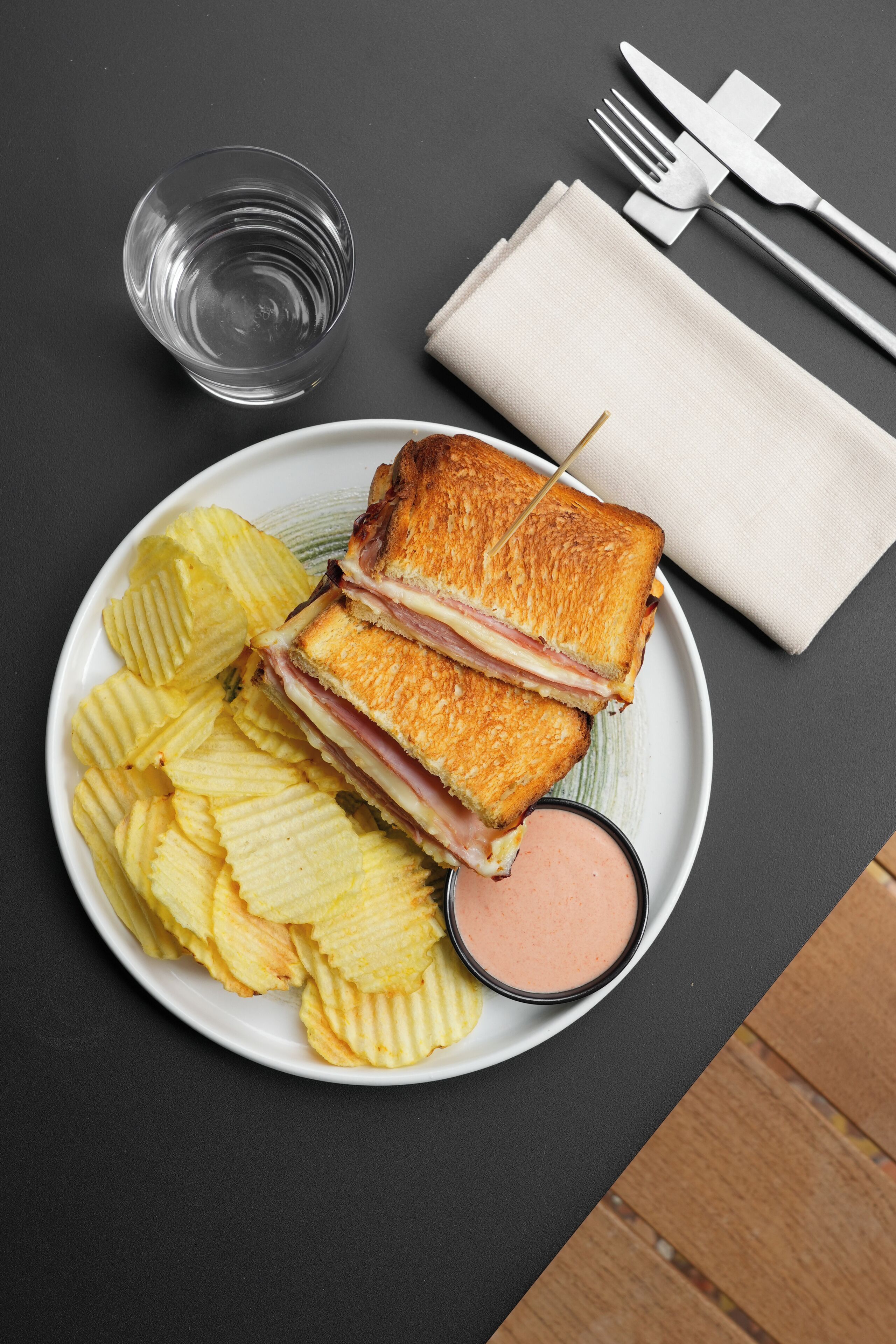 Een bord met een gegrilde sandwich, chips en een klein bakje saus.