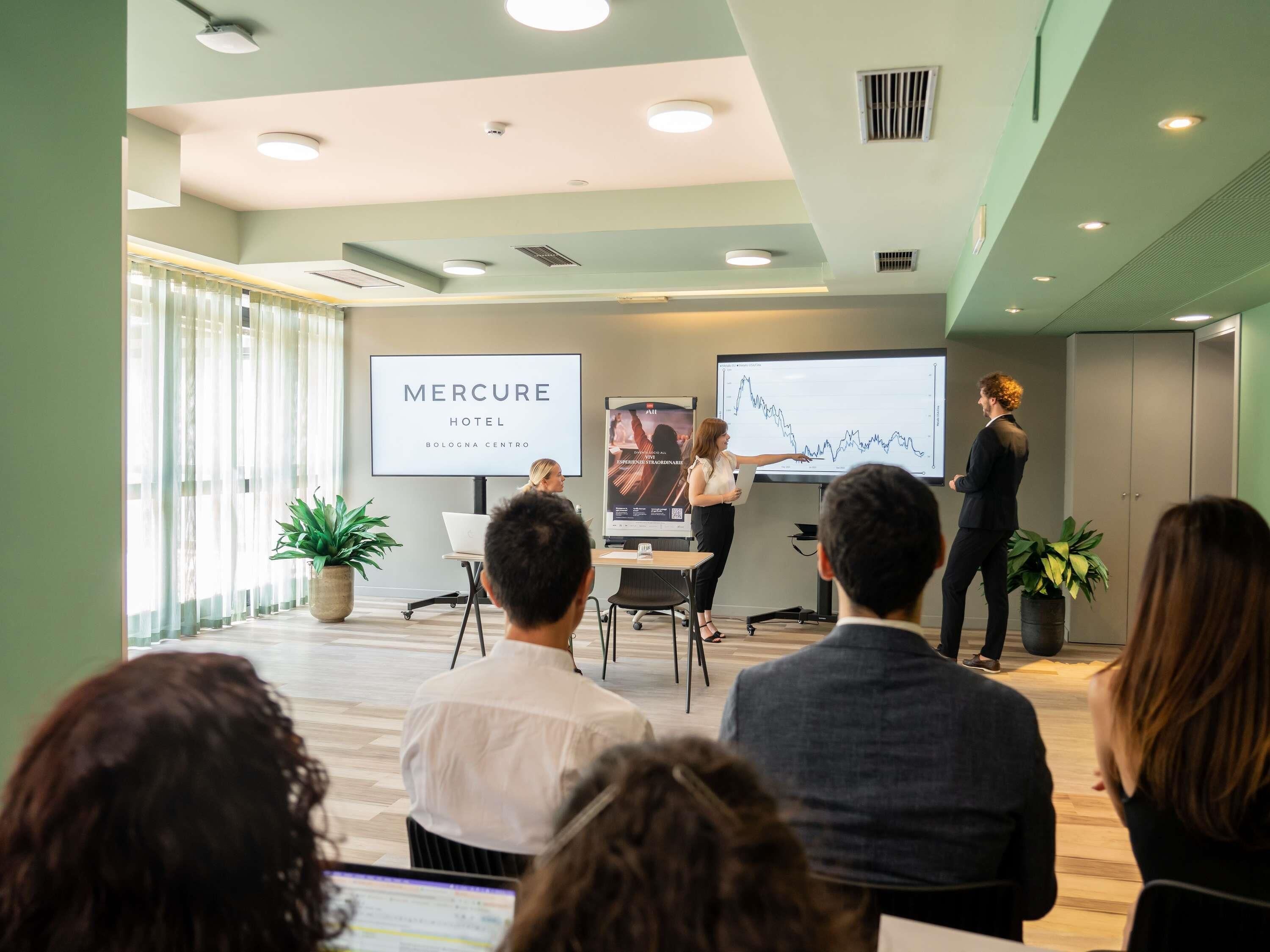 Una sala conferenze con uno schermo per presentazioni che mostra un grafico e il testo 'MERCURE HOTEL BOLOGNA CENTRO'.
