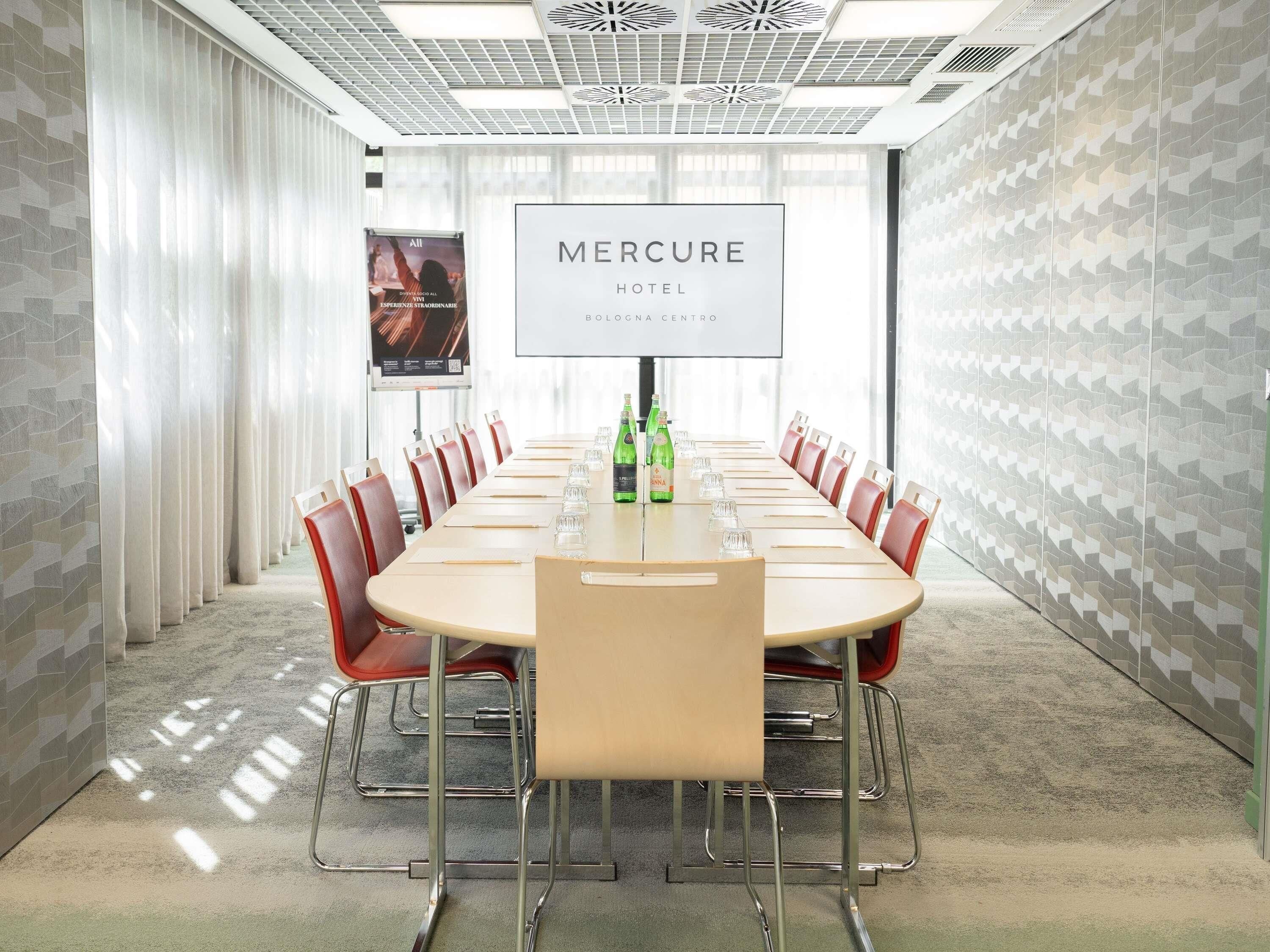 Un salón de conferencias con una larga mesa, sillas rojas y una pantalla grande que muestra el nombre del hotel.
