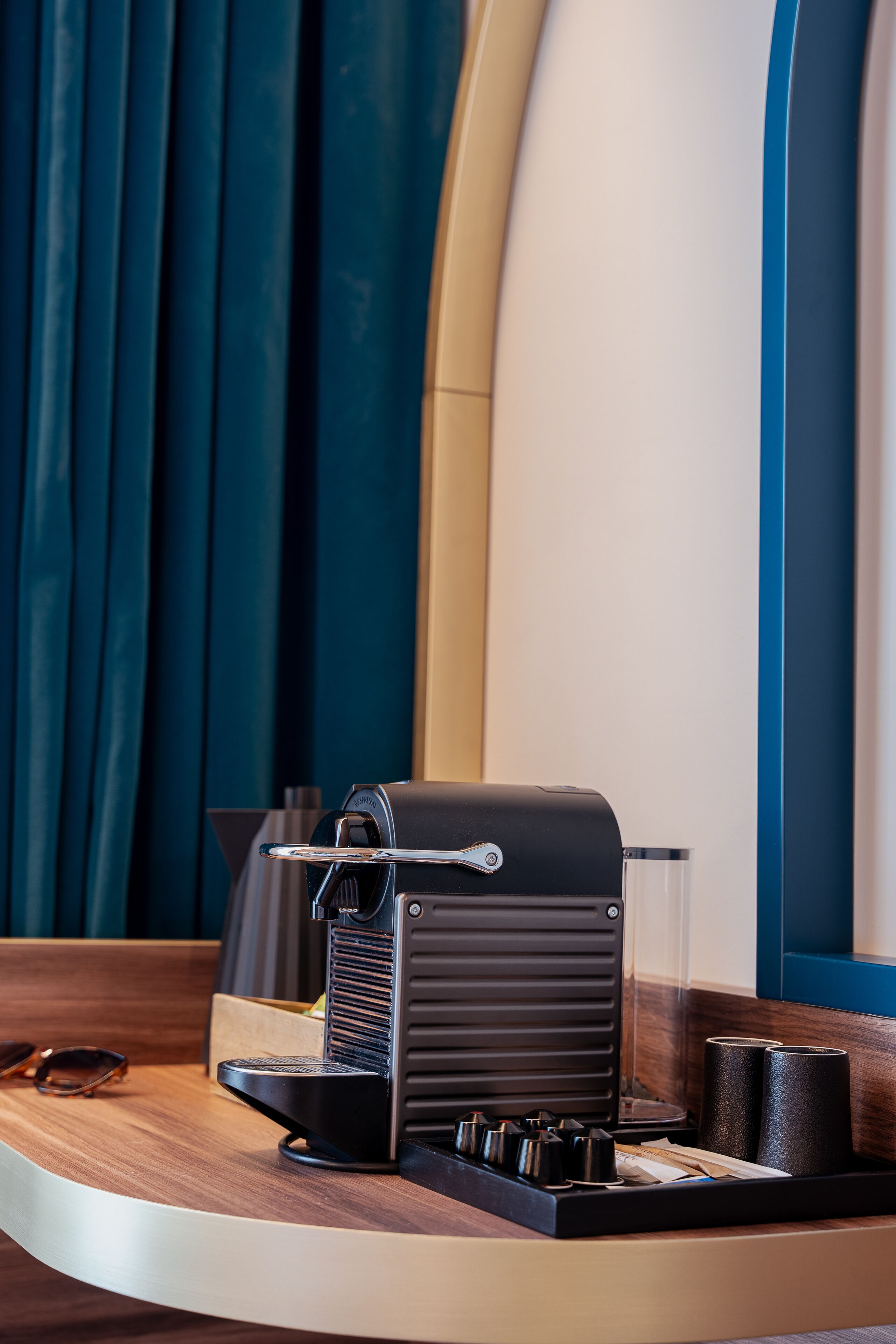 A coffee machine on a wooden counter with a glass cup, a pair of sunglasses, and a notebook.