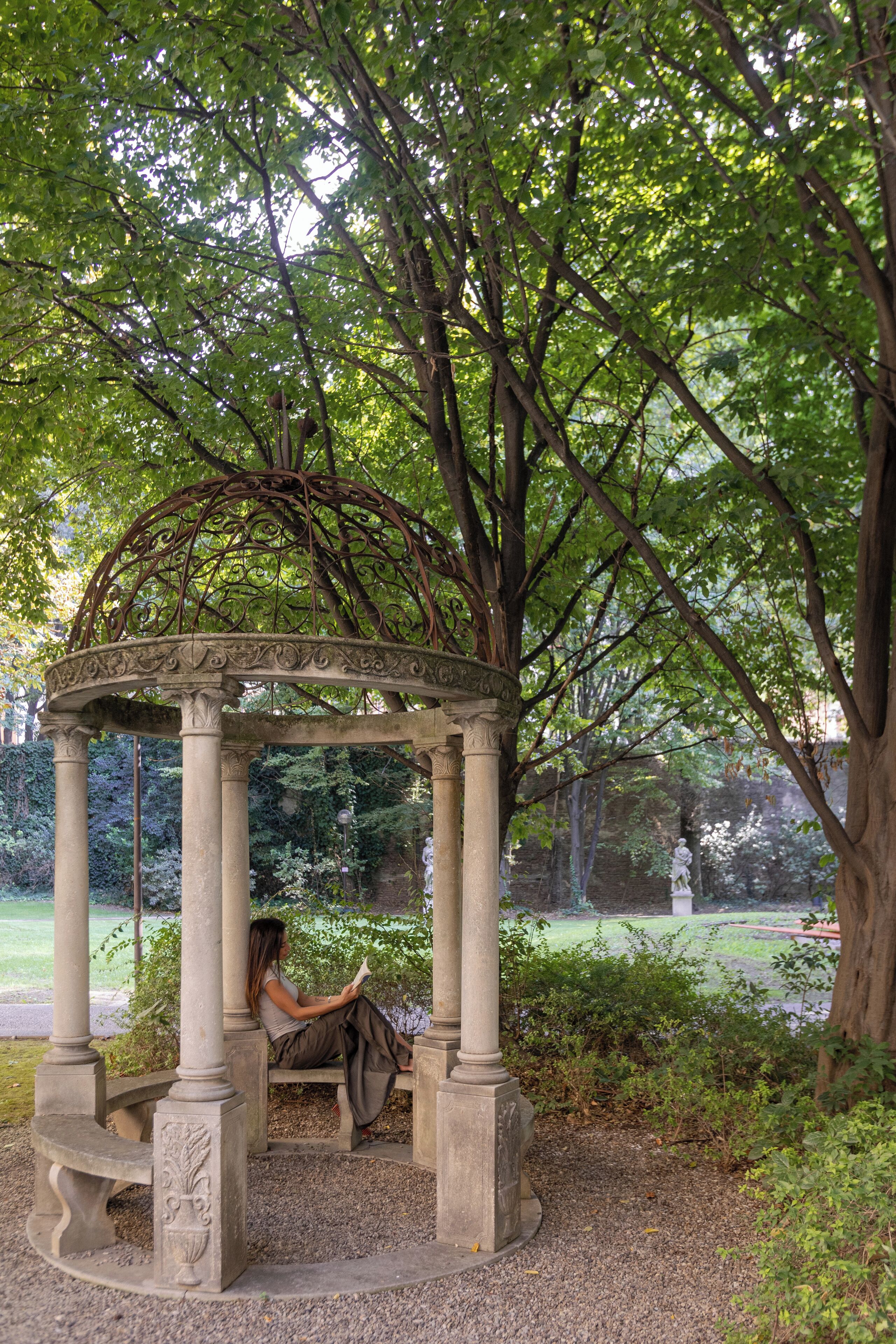 A gazebo with a person reading a book, surrounded by greenery and trees.