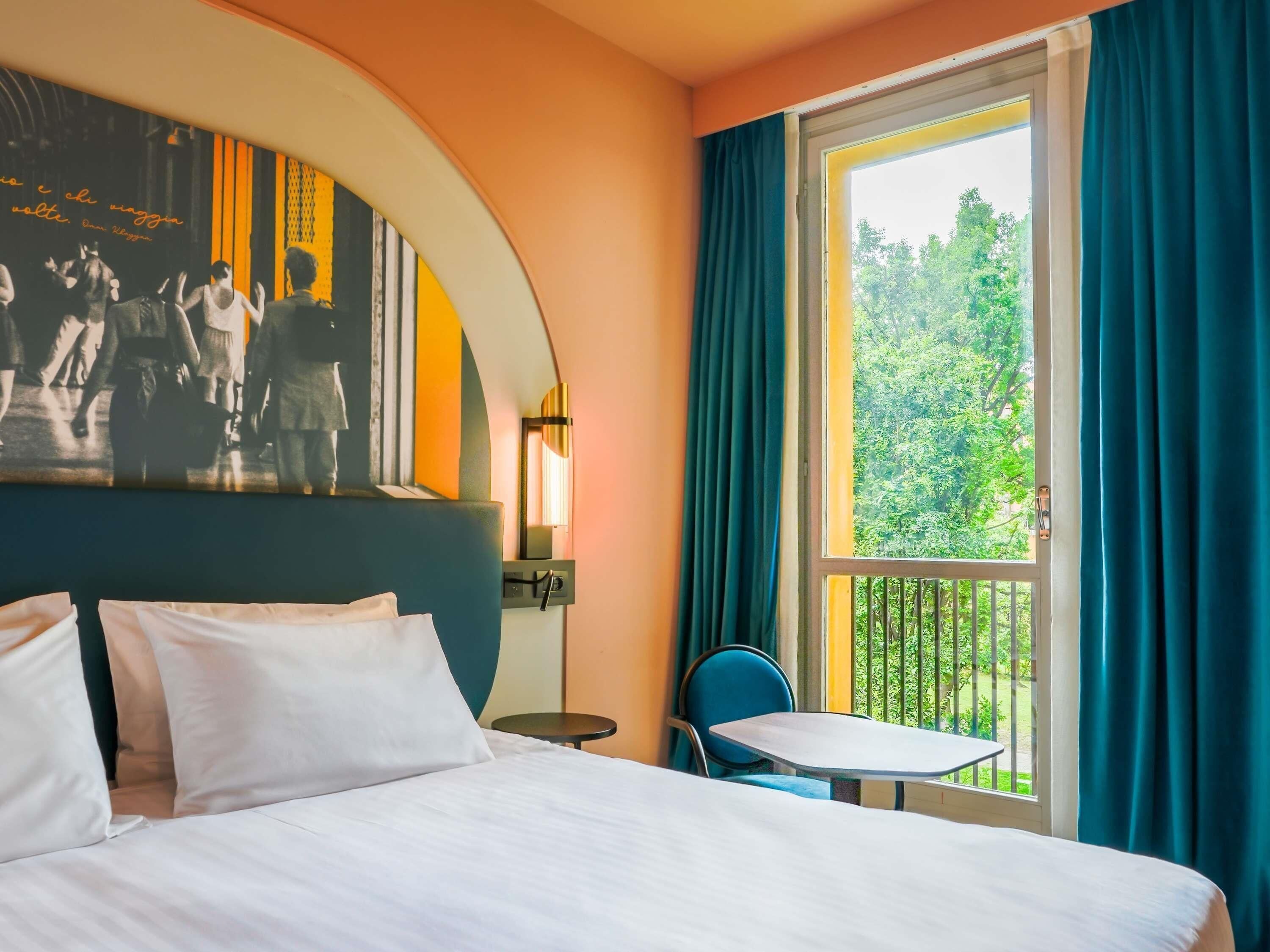 A hotel room with a bed, a small table, a chair, a window with curtains, and a wall-mounted light.
