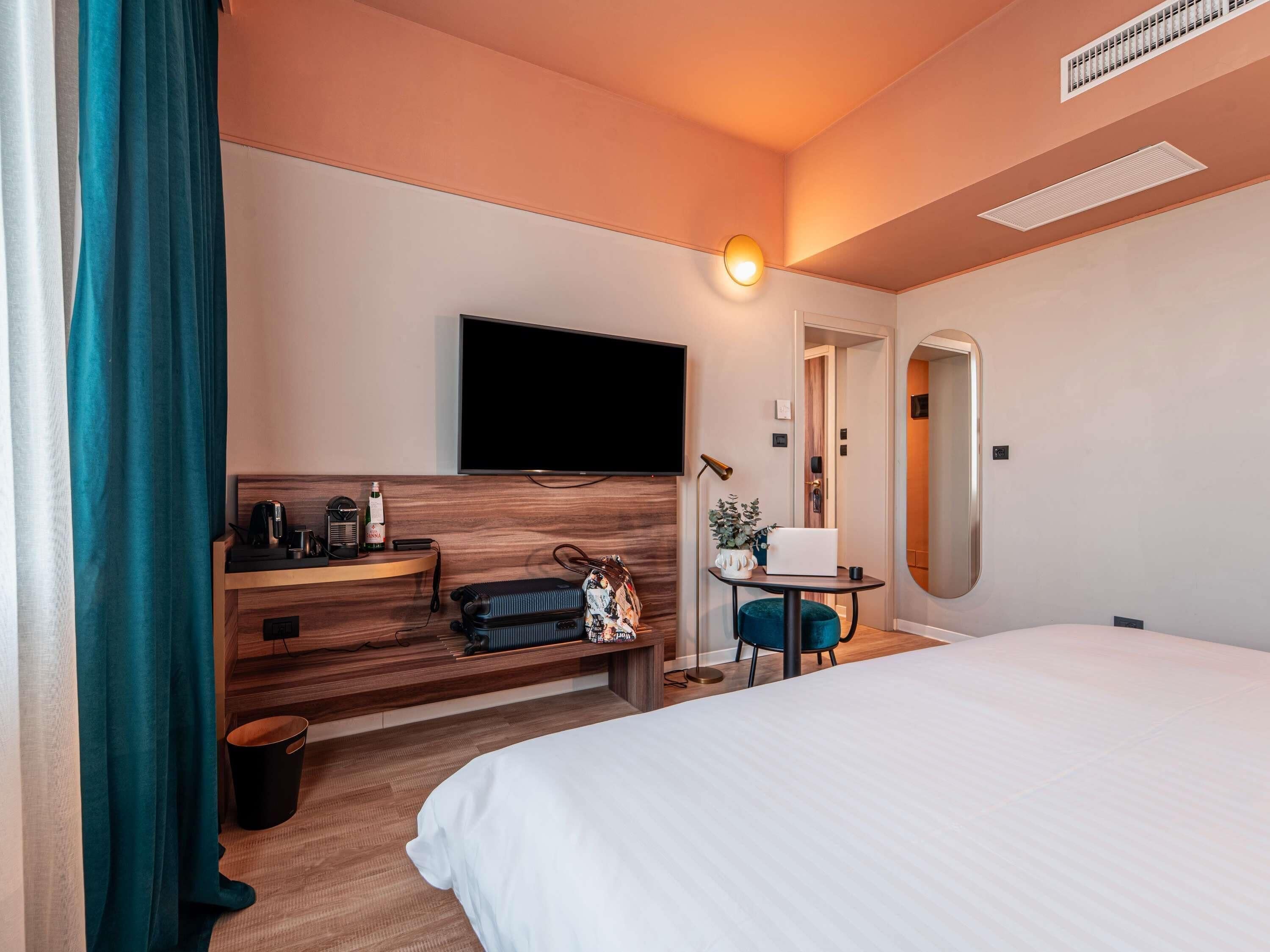 A hotel room with a bed, a desk with a flat-screen TV, a small table with a plant, and a mirror on the wall.