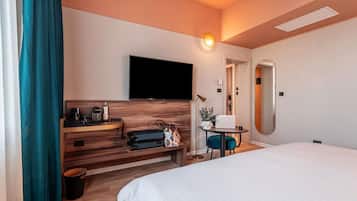 Una habitación de hotel con una cama, un escritorio con televisor de pantalla plana, una pequeña mesa con una planta y un espejo en la pared.