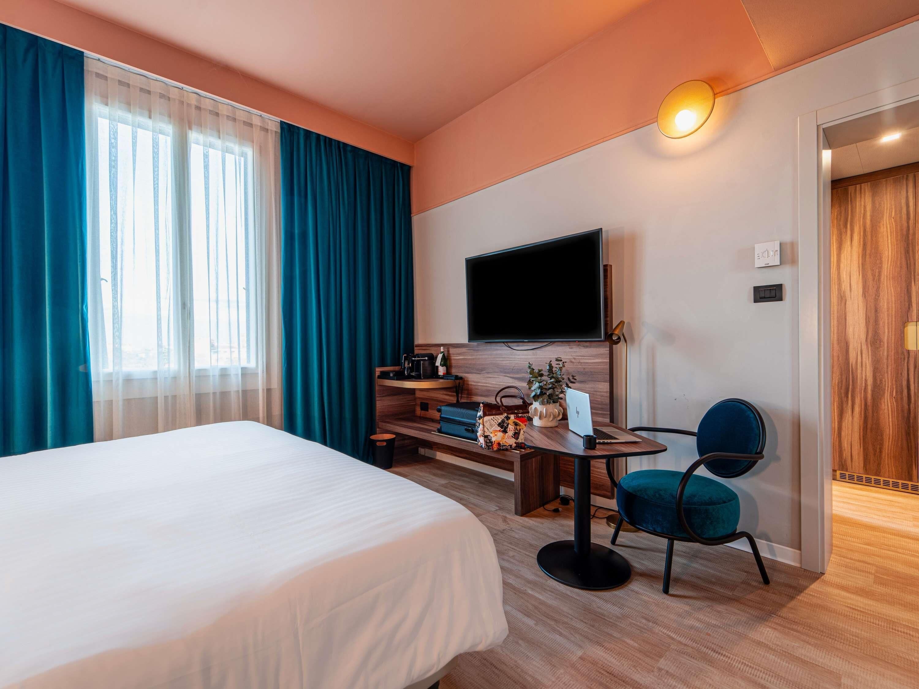 Habitación de hotel con una cama, un escritorio con una computadora portátil, una silla, un televisor y una puerta.