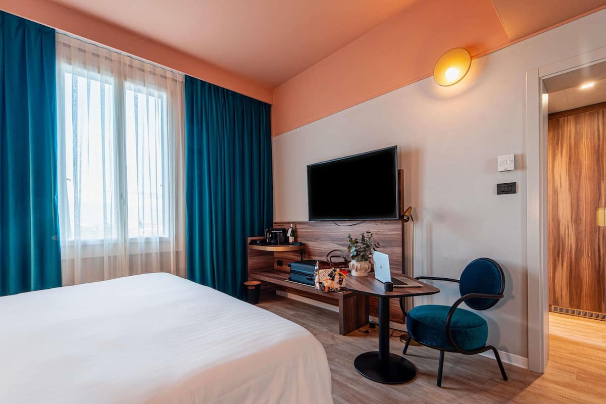 Ein Hotelzimmer mit einem Bett, einem Schreibtisch mit einem Laptop, einem Sessel, einem Fernseher und einer Tür.