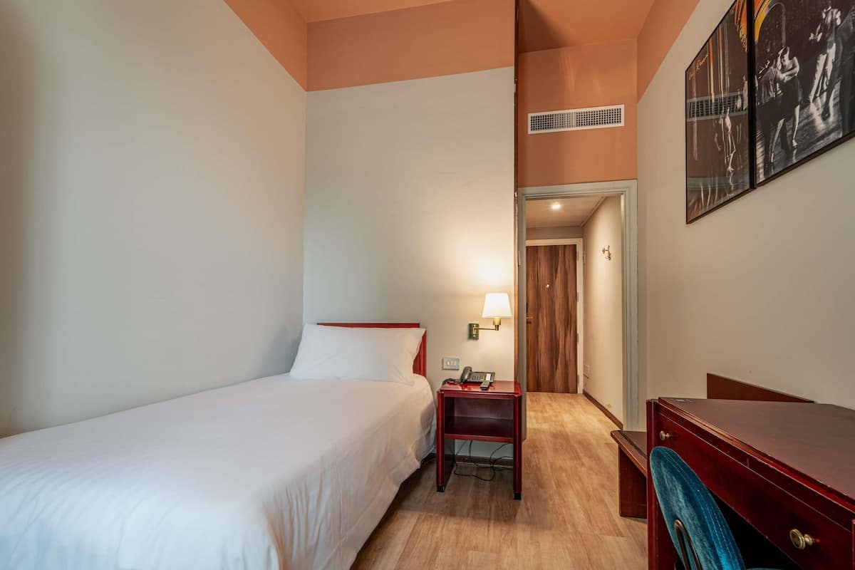 Ein Hotelzimmer mit einem Bett, einem Schreibtisch, einem Stuhl, einer Lampe und einer Tür zu einem anderen Raum.