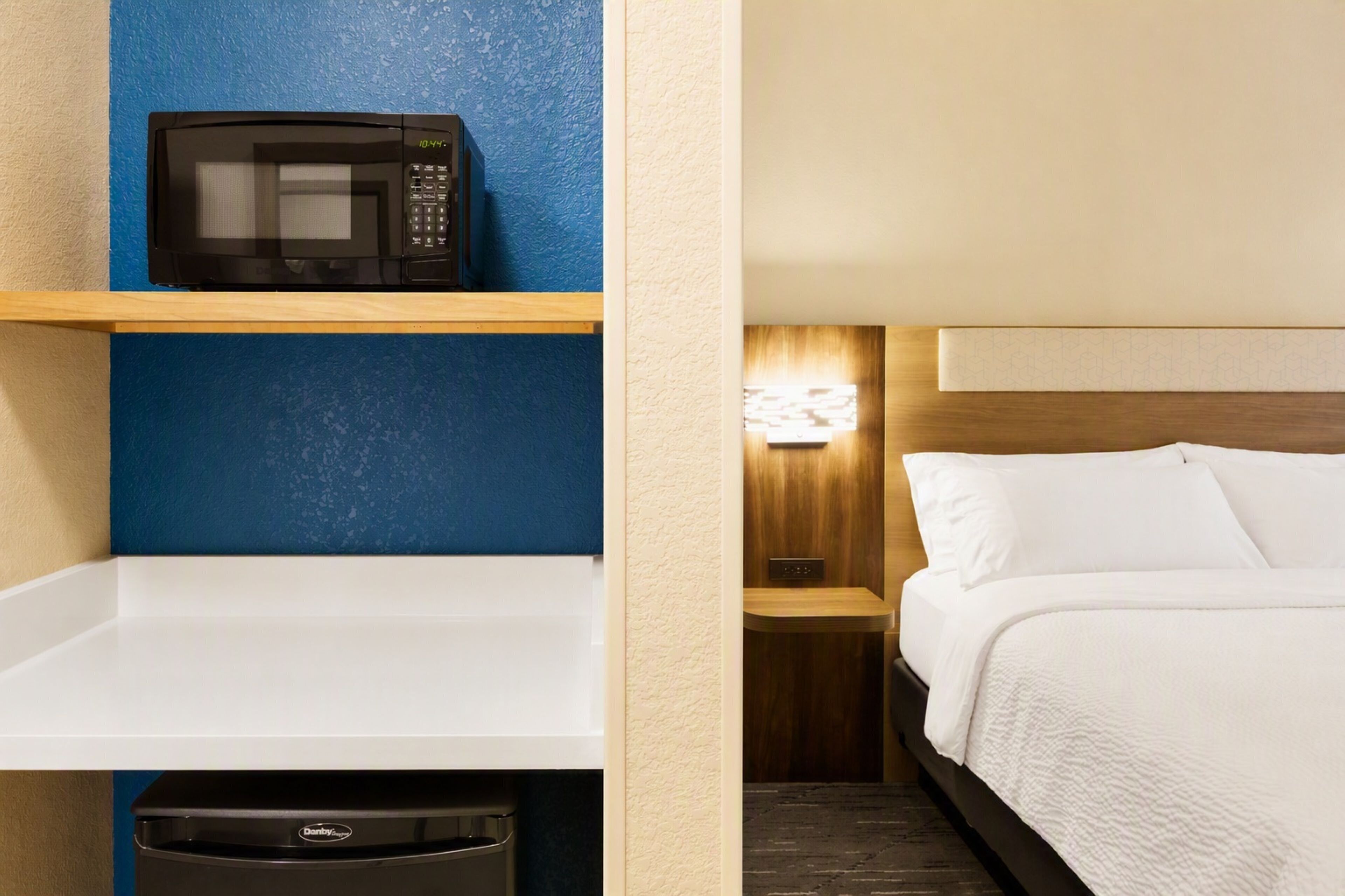 A hotel room with a microwave on a shelf, a bed with white bedding, and a nightstand with a lamp.