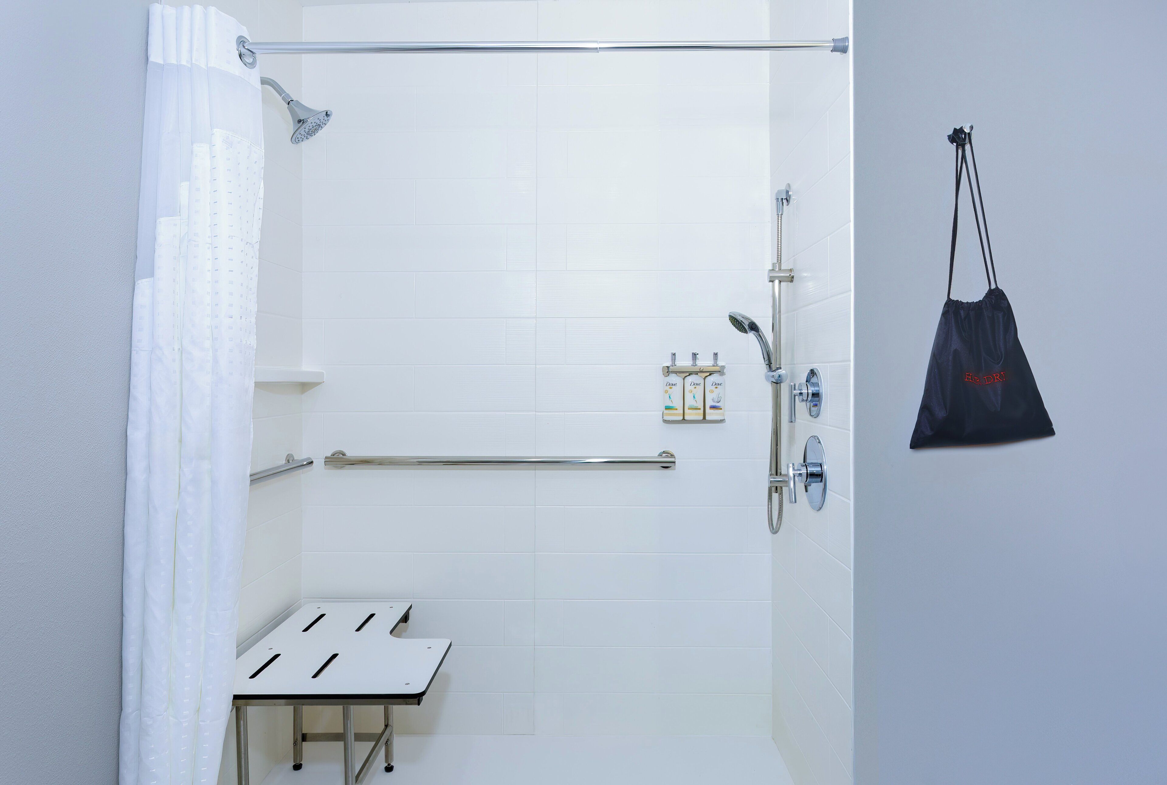 A modern bathroom with white tiles, a shower with a handheld showerhead, a grab bar, and a small table.