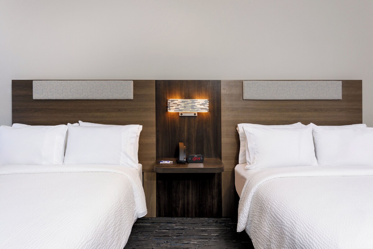 A hotel room with two beds, a nightstand with a clock, and a wall-mounted light fixture.