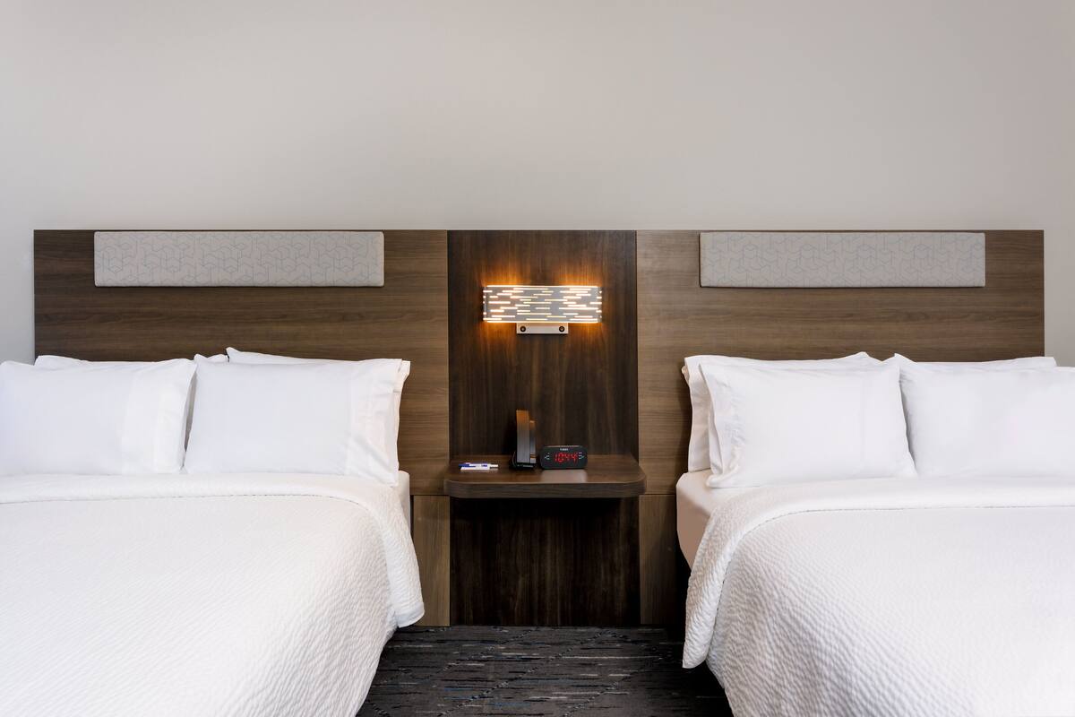 Hotellihuone, jossa on kaksi sänkyä, yöpöytä, jossa on kello, ja seinään kiinnitetty valaisin.