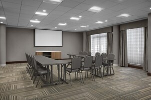 Uma sala de conferências com tela de projeção, uma mesa comprida com cadeiras e janelas grandes com cortinas.