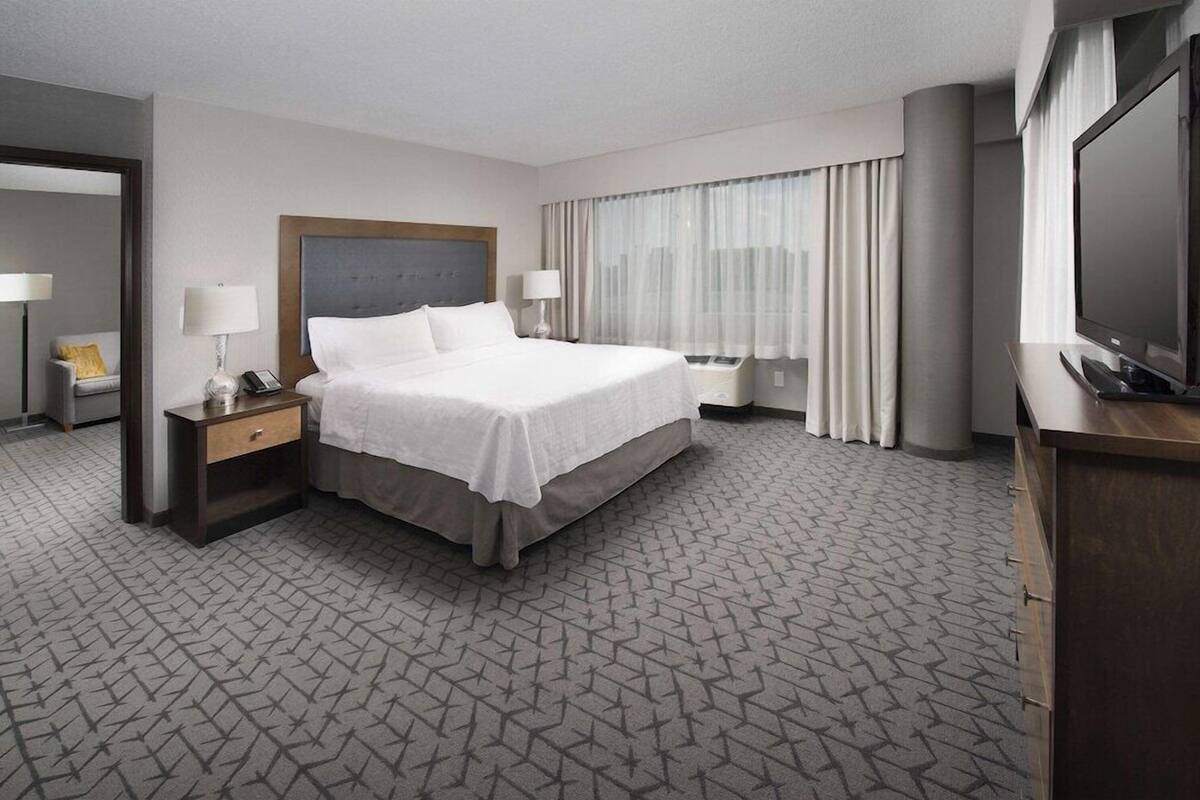 A hotel room with a large bed, bedside tables with lamps, a television, and a view of the city through the window.