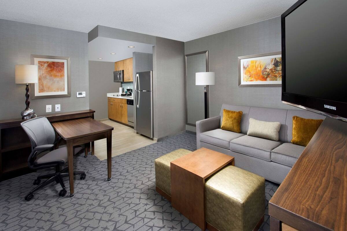 A hotel room with a dining area, kitchenette, grey sofa, and a TV.