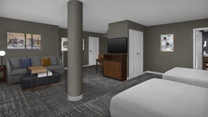 Habitación de hotel con alfombra gris, un sofá, un televisor, un aparador y dos camas.