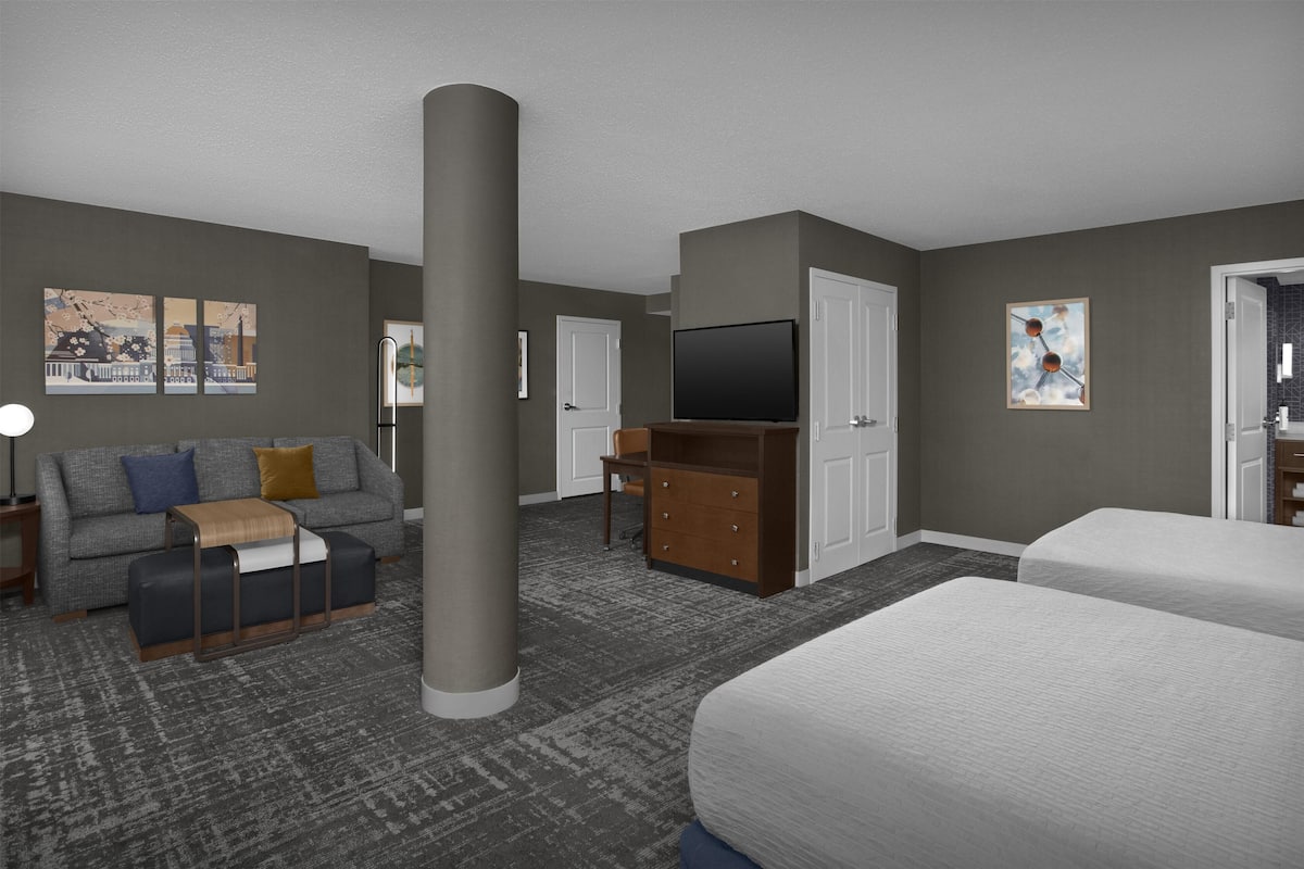 A hotel room with a grey carpet, a sofa, a TV, a dresser, and two beds.