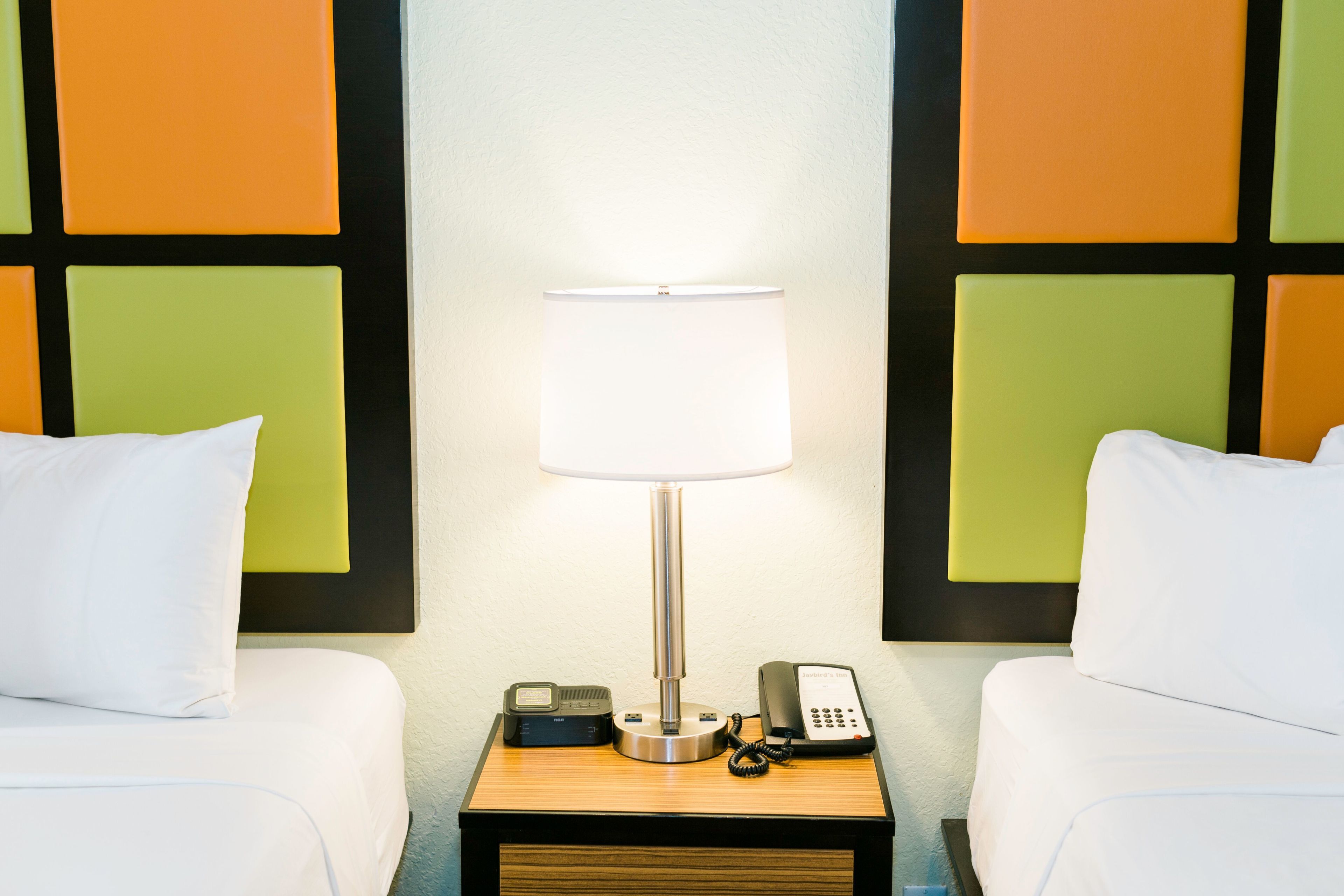 A hotel room with two beds, a bedside table with a lamp, a telephone, and a small device on the table.