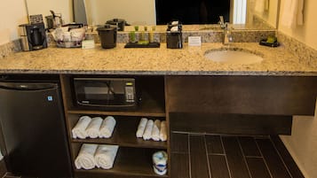 A hotel bathroom with a granite countertop, a sink, a microwave, and shelves with towels and toiletries.