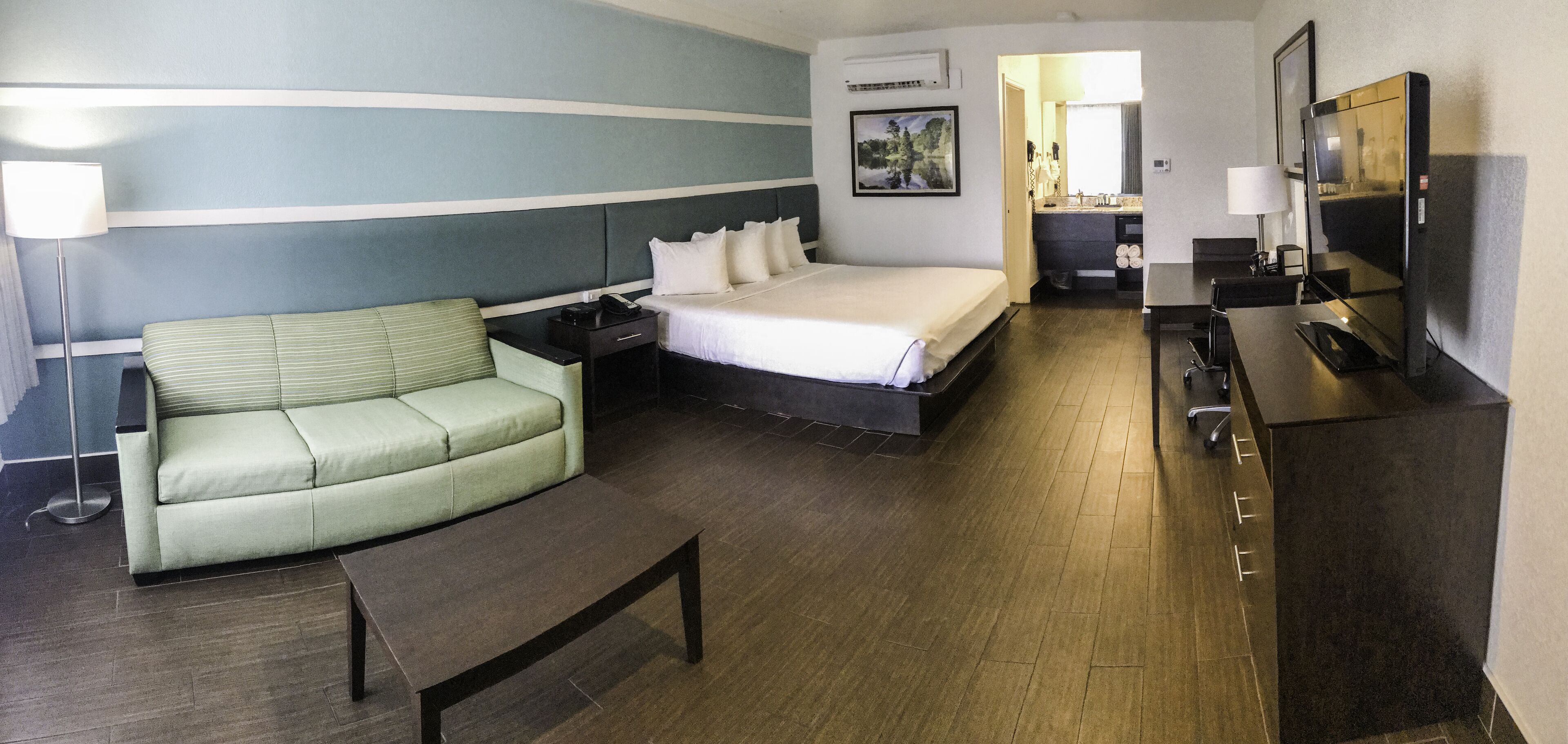 A hotel room with a bed, a sofa, a desk, a TV, and a bathroom visible through an open door.