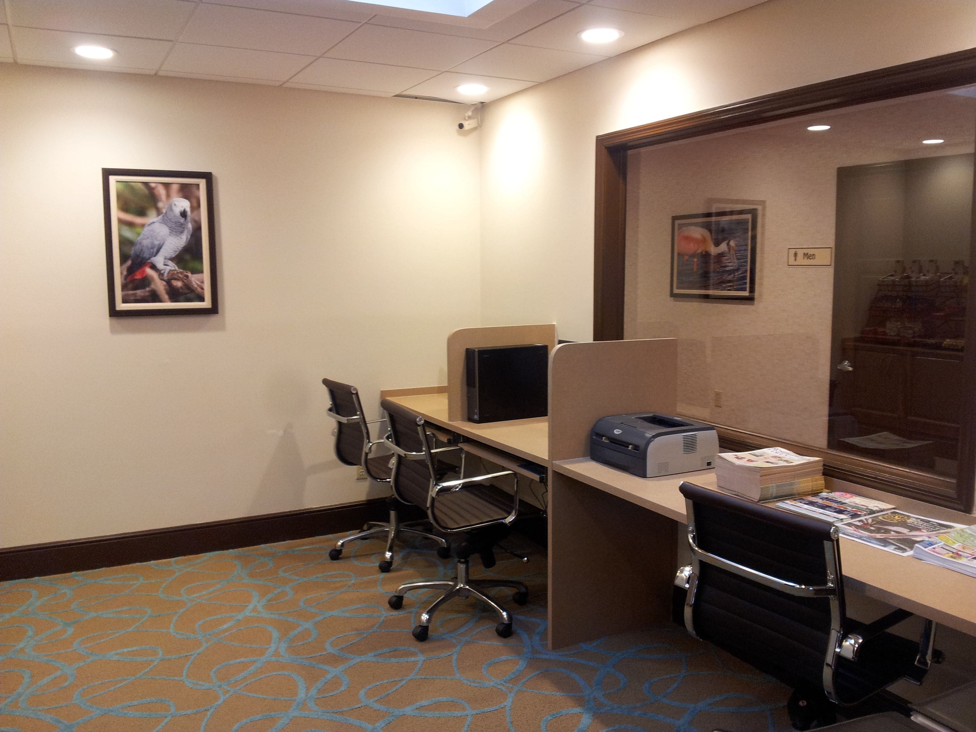 A hotel room with a desk, chairs, a printer, and a framed picture on the wall.