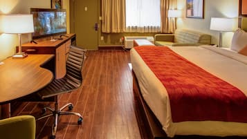 A hotel room with a bed, a desk with a computer, a chair, a TV, and a window with curtains.