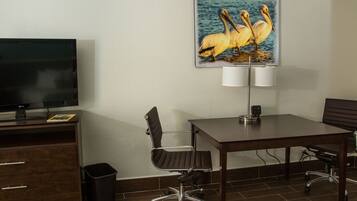 A hotel room with a flat-screen TV, a painting of pelicans, a desk, two chairs, a lamp, and a trash bin.