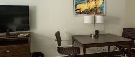 A hotel room with a flat-screen TV, a painting of pelicans, a desk, two chairs, a lamp, and a trash bin.