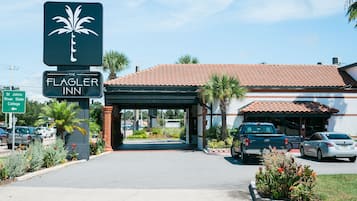 The exterior of the Flagler Inn, featuring a palm tree logo, a covered entrance, and a parking area with cars.